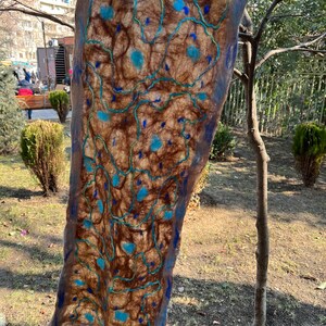 Könnte beinhalten: Ein brauner und blauer Filzschal mit einem einzigartigen Design. Der Schal hat eine braune Basis mit blauen Akzenten und einem blauen Rand. Das Design umfasst blaue Kreise und Linien, die ein strukturiertes Aussehen erzeugen. Der Schal wird im Freien präsentiert.