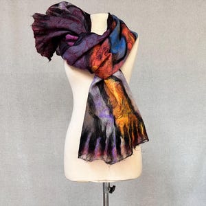 Puede incluir: Bufanda de fieltro colorida drapeada sobre un maniquí. La bufanda presenta una mezcla de colores, incluyendo morado oscuro, naranja, azul y negro. La textura de la bufanda parece suave y fluida, con un diseño artístico único.