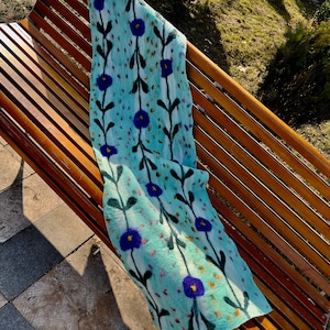 Könnte beinhalten: Ein hellblauer Schal mit einem Blumenmuster aus dunkelblauen Blüten und schwarzen Stielen. Der Schal besteht aus einem weichen Material und ist über einer Holzbank drapiert. Das Blumenmuster wiederholt sich über den Schal.