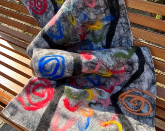May include: A gray felt scarf with colorful embroidered spirals and lines. The scarf has black accents and is folded on a wooden bench. The embroidery includes red, orange, yellow, green, blue, and pink threads.
