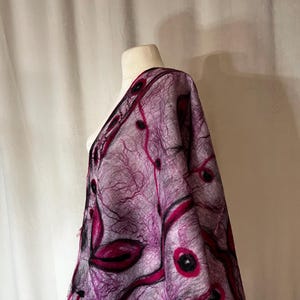 Könnte beinhalten: Ein Filzschal aus Wolle, der über einer Schaufensterpuppe drapiert ist. Der Schal hat ein komplexes Design mit Schattierungen von Lila, Schwarz und Magenta. Das Muster umfasst organische Formen und Linien, wodurch ein strukturiertes und künstlerisches Aussehen entsteht. Der Hintergrund ist schlicht weiß.