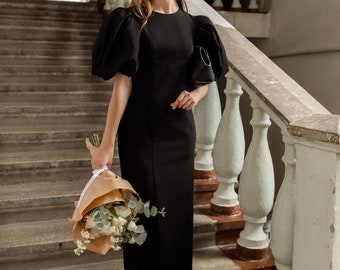 May include: A woman in a black dress with large puffed sleeves stands on a staircase holding a bouquet of white flowers wrapped in brown paper.