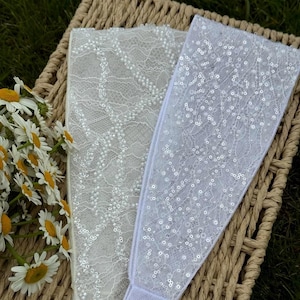 Peut inclure: Deux bandeaux blancs avec des éléments décoratifs reposent sur un panier en osier. L'un présente un motif en dentelle avec de petites perles. L'autre est orné de paillettes. Un bouquet de marguerites aux centres jaunes et aux pétales blancs est dans le coin.