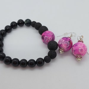 May include: A black and lava rock bracelet with a pink and white patterned bead, and matching pink and white patterned earrings with silver accents.
