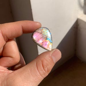 May include: A teardrop-shaped labradorite gemstone with a polished surface. The stone displays a vibrant play of color, with hues of pink, purple, blue, and gold. The gemstone is held between fingers.