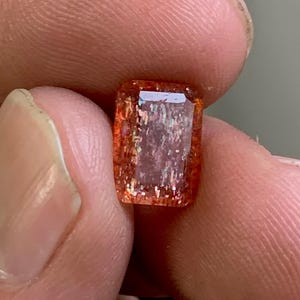 Könnte beinhalten: Ein rechteckiger, orangefarbener Edelstein mit Facettenschliff. Der Edelstein hat eine reflektierende, irisierende Qualität, mit Farbflecken, die in seinen Tiefen sichtbar sind. Der Stein wird zwischen den Fingern gehalten, was seine Größe und Klarheit zeigt.