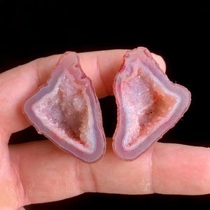 May include: Two pink and white geode slices with a crystalline interior. The outer edges are a darker pink, with a white border. The center of each geode is filled with sparkling crystals in shades of pink and beige. The background is black.