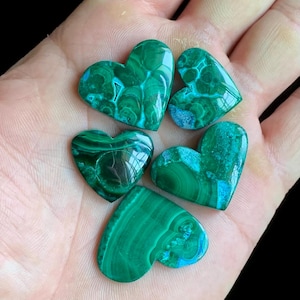 Könnte beinhalten: Fünf herzförmige Malachitsteine mit verschiedenen Grün- und Blaumustern. Die polierten Steine haben eine glänzende Oberfläche, die die natürlichen Maserungen und Farbvariationen hervorhebt. Die Herzen werden vor einem neutralen Hintergrund präsentiert.