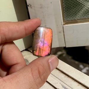 Könnte beinhalten: Ein rechteckiger, polierter Labradorit-Edelstein mit einem lebendigen Farbspiel in Rosa, Orange und Lila. Der Stein wird zwischen Fingern gehalten, wodurch sein Farbspiel und seine natürlichen Muster zur Geltung kommen. Die Oberfläche reflektiert das Licht.
