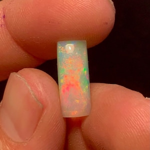 May include: A rectangular opal gemstone with a polished surface. The opal displays a play of color, with flashes of red, orange, green, and yellow. The gemstone is held between two fingers against a dark background.
