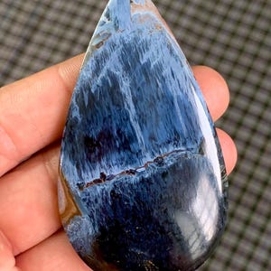 May include: A polished, teardrop-shaped gemstone with a striking blue and black pattern. The stone features swirling, textured patterns in shades of deep blue, black, and hints of brown. The gemstone is held in a hand, showcasing its smooth, glossy surface.