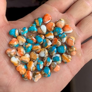 Könnte beinhalten: Eine Sammlung herzförmiger Perlen in den Farbtönen Orange, Türkis, Weiß und Gold. Die Perlen sind klein und scheinen aus einem Verbundmaterial zu bestehen. Die Perlen werden in der Hand einer Person präsentiert.