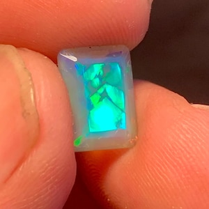 May include: A rectangular opal gemstone with a vibrant display of green and blue internal colors. The gem is held between two fingers, showcasing its polished surface and unique color play. The opal's shape is a rectangle.