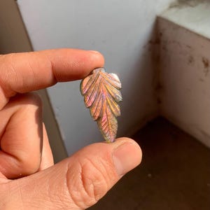 Könnte beinhalten: Ein geschnitzter Blattanhänger mit schillernden rosa, lila und orangefarbenen Farbtönen. Der Edelstein wird zwischen den Fingern gehalten und zeigt die komplizierten Details der Blattadern und -kanten. Der Anhänger ist ein einzigartiges Schmuckstück.