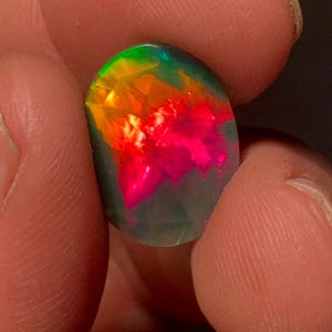 Könnte beinhalten: Ein ovaler schwarzer Opal-Edelstein mit einer dunklen Grundfarbe und leuchtenden Blitzen in Neonrot, Orange und Grün. Die Oberfläche des Steins ist facettiert, wodurch ein geometrisches Muster entsteht, das das Licht auf der Oberfläche reflektiert.