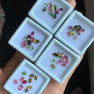 Puede incluir: Surtido de gemas de colores en peque&ntilde;as cajas de exhibici&oacute;n blancas. Las gemas son de varias formas y tama&ntilde;os, incluyendo cortes ovalados, en forma de pera y redondos. Los colores incluyen rosa, verde, marr&oacute;n y amarillo. Las gemas se muestran en una mano.