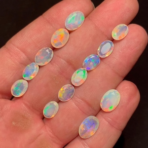 Puede incluir: Una colección de ópalos ovalados, cada uno mostrando un juego de color único con destellos de naranja, verde y azul. Las piedras preciosas se sostienen en una mano, mostrando sus cualidades iridiscentes. Los ópalos varían en tamaño y brillo.