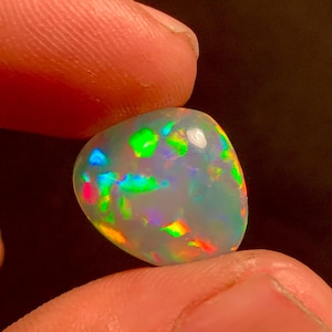 Könnte beinhalten: Ein weißer Opal-Edelstein in Tropfenform mit irisierenden Schimmern in Grün, Blau, Rot und Orange.