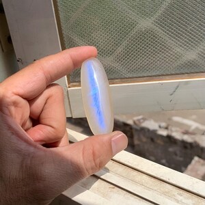 Könnte beinhalten: Ein ovaler Mondstein-Cabochon mit einer glatten, durchscheinenden Oberfläche und einem ausgeprägten, zentrierten blauen Schimmer. Der Edelstein ist länglich und poliert und zeigt eine weiche weiße Grundfarbe mit einem hellen, vertikalen blauen Lichtreflex.