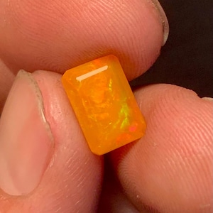 Könnte beinhalten: Ein rechteckiger, orangefarbener Edelstein mit hellem, innerem Leuchten. Der Edelstein wird zwischen zwei Fingern gehalten, wodurch seine polierten Facetten und seine leuchtende Farbe zur Geltung kommen. Der Stein hat einen leichten gelben und roten inneren Blitz.
