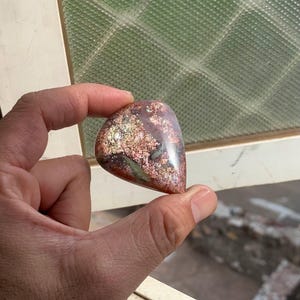 May include: A polished, teardrop-shaped stone featuring a complex mix of metallic copper, bronze, and deep red hues with shimmering, reflective inclusions throughout its smooth, rounded surface, displaying a natural, multi-coloured mineral pattern.