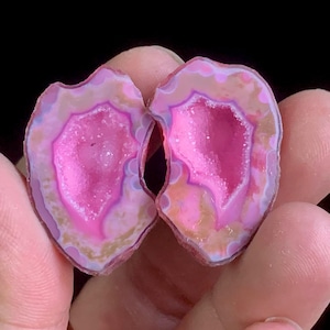 Könnte beinhalten: Zwei Hälften einer Geode, die einen leuchtend rosa Kristallkern zeigen. Die äußeren Ränder weisen Bänder aus hellrosa, beige und violett auf. Die Geode wird zwischen Fingern vor dunklem Hintergrund gehalten.