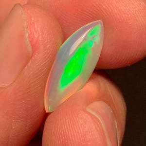 Könnte beinhalten: Ein marquiseförmiger Opal-Edelstein mit einer durchscheinenden, milchig-weißen Basis und einem lebendigen, länglichen Blitz in Neongrün, der durch die Mitte verläuft. Der Stein hat eine glatte, polierte Oberfläche und ein spitz zulaufendes Design.
