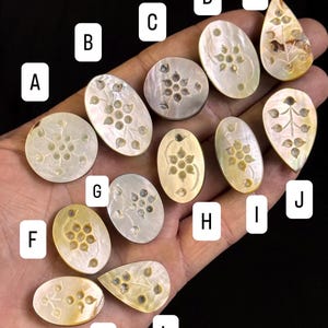 Könnte beinhalten: Eine Sammlung von Perlmutt-Cabochons in verschiedenen Formen, darunter oval, rund und tropfenförmig. Jeder Cabochon weist ein eingraviertes Blumenmuster auf. Die Farben reichen von cremeweiß bis goldgelb, mit dezentem Schimmer.