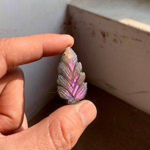 Könnte beinhalten: Ein geschnitzter Edelstein in Form eines Blattes, gehalten zwischen Fingern. Der Stein hat eine graue Grundfarbe mit schillernden rosa und lila Farbtönen. Das Blattdesign weist detaillierte Adern und eine strukturierte Oberfläche auf, die das Licht einfängt.