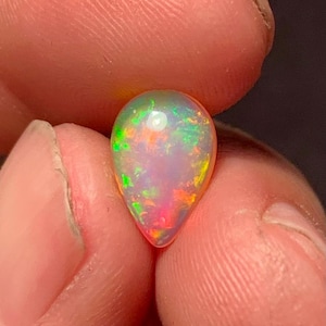Könnte beinhalten: Ein tropfenförmiger Opal-Edelstein mit einer lebendigen Farbpalette, darunter Grün, Orange und Rot, gehalten zwischen Fingern. Der Edelstein hat eine milchig-weiße Basis mit Farbflecken.