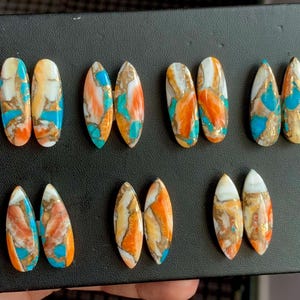 Könnte beinhalten: Eine Sammlung ovaler und tropfenförmiger Cabochon-Edelsteine. Die Steine weisen eine Mischung aus Farben auf, darunter Türkis, Orange, Weiß und Gold, mit marmoriertem oder gemustertem Aussehen. Die Edelsteine werden auf einer schwarzen Oberfläche präsentiert.