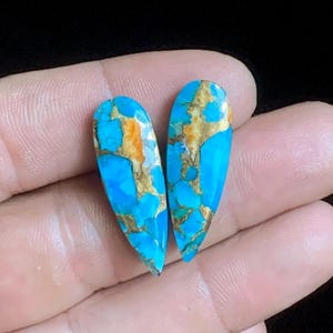 May include: Two teardrop-shaped gemstones with a vibrant turquoise hue, accented by intricate patterns of gold and brown. The stones are held in a hand, showcasing their polished surface and unique color variations. The gemstones are likely for jewelry making.