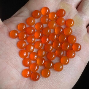 May include: A collection of polished, round, orange gemstones. The gemstones are a vibrant, translucent orange colour and appear to be cabochons. The stones are displayed in a hand, with a dark background.