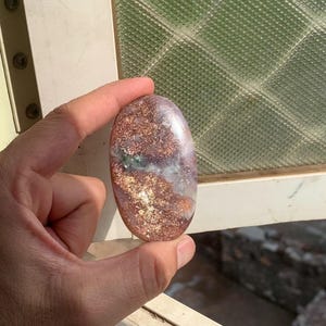 Könnte beinhalten: Ein ovaler, polierter Edelstein mit einer Mischung aus kupferfarbenen metallischen Einschlüssen, durchscheinenden lavendelfarbenen Bereichen und kleinen Flecken aus dunkelgrünen Mineralablagerungen mit einer glatten, reflektierenden Oberfläche.