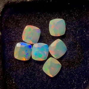 May include: Six square-cut opals with iridescent color flashes of blue, green, orange, and yellow. The gemstones are set against a dark, textured background, showcasing their brilliance. Each opal has a unique play of color.