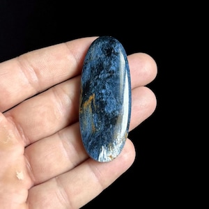 Könnte beinhalten: Ein oval geformter Edelstein mit polierter Oberfläche. Der Stein weist ein wirbelndes Muster aus tiefem Blau, Schwarz und Hauch von Gold und Weiß auf. Der Edelstein wird in einer Hand vor schwarzem Hintergrund gehalten.