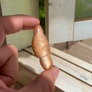 Könnte beinhalten: Ein polierter, goldbrauner Edelstein mit einer einzigartigen, länglichen Form. Der Stein hat eine schimmernde, strukturierte Oberfläche und wird zwischen zwei Fingern gehalten. Die Farbe des Edelsteins reicht von hell- bis dunkelbraun, mit einem subtilen Glitzern.