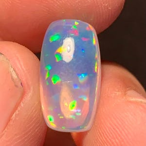 Könnte beinhalten: Ein polierter, rechteckiger Opal-Edelstein mit einer durchscheinenden, milchig-weißen Basis. Der Opal zeigt lebhafte Farbblitze, darunter Grün, Blau und Rot, wodurch ein Farbspiel-Effekt entsteht. Der Edelstein wird zwischen zwei Fingern gehalten.
