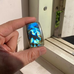 Könnte beinhalten: Ein rechteckiger Labradorit-Edelstein mit polierter Oberfläche. Der Stein zeigt schillernde blaue, grüne und goldene Reflexe. Der Edelstein wird vor einem weißen Hintergrund gehalten, um seine leuchtenden Farben und natürlichen Muster zu präsentieren.