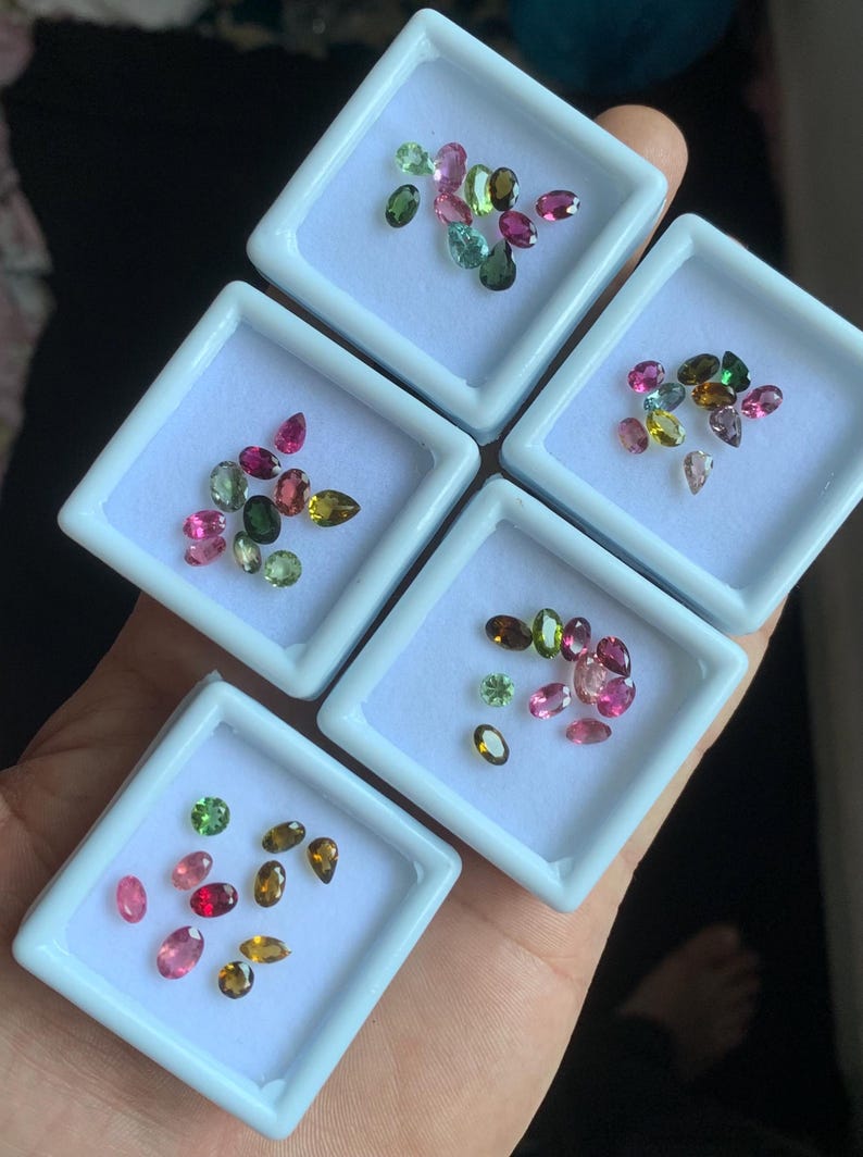 Puede incluir: Un surtido de piedras preciosas de turmalina coloridas en varias formas y tama&ntilde;os, incluyendo cortes ovalados, en forma de pera y redondos. Las piedras preciosas se exhiben en peque&ntilde;os recipientes cuadrados blancos, mostrando una gama de tonos rosas, verdes y marrones.