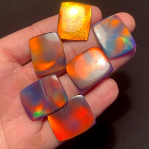 May include: Six iridescent, square gemstones in a hand. The stones show orange, purple, blue, and green hues, with a polished finish. One stone is a solid gold colour. The gemstones are approximately 2 cm square.