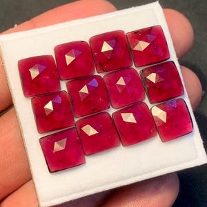 May include: A collection of eleven square-cut, faceted, deep magenta-red gemstones arranged in a grid pattern on a white display card. Each gemstone features a distinct rose-cut top with a central triangular facet that reflects light brightly.