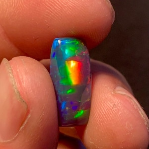 Könnte beinhalten: Ein rechteckiger schwarzer Opal-Edelstein mit lebendiger Regenbogen-Irisierung. Der polierte Stein zeigt Blitze aus blauem, grünem und rotem Licht. Der Opal wird zwischen zwei Fingern gehalten und zeigt sein Farbspiel.