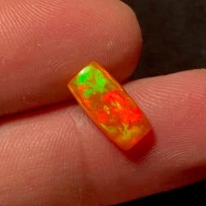 Könnte beinhalten: Ein rechteckiger Opal-Edelstein mit leuchtenden Farben wie Orange, Rot und Grün. Der Edelstein wird zwischen zwei Fingern gehalten, was seine polierte Oberfläche und das innere Farbspiel hervorhebt. Die Abmessungen des Opals sind nicht sichtbar.