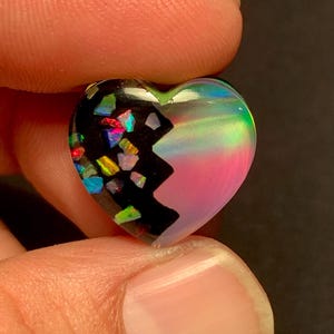 Könnte beinhalten: Ein herzförmiger Cabochon mit geteiltem Design. Eine Seite ist schwarz mit eingebetteten, schillernden, bunten Flecken in Rot, Blau, Grün und Gelb. Die andere Seite besteht aus einem glatten, durchscheinenden Material mit sanftem rosa und grünem Schimmer.