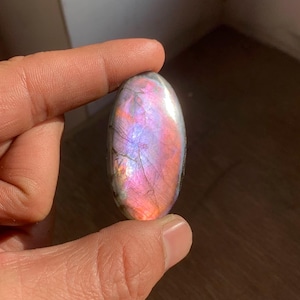 Puede incluir: Una piedra preciosa de labradorita de forma ovalada con una superficie pulida. La piedra muestra un vibrante juego de colores, con tonos de rosa, morado y azul. La gema se sostiene entre dos dedos.