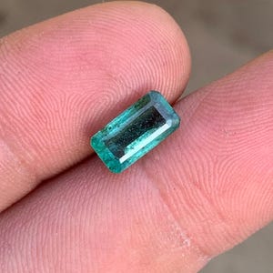 May include: A rectangular emerald gemstone with a faceted cut, displaying a deep green color with natural internal inclusions and a polished surface, held between two fingers to showcase its size and translucent quality.