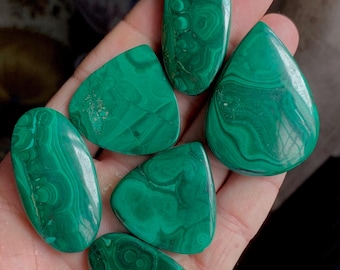 May include: A collection of polished malachite gemstones in various shapes, including oval, teardrop, and triangular forms. The stones display a vibrant green color with intricate swirling patterns, showcasing natural mineral formations. The gemstones are held in a hand.