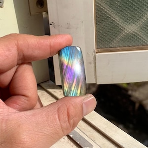 Könnte beinhalten: Ein rechteckiger Labradorit-Edelstein-Cabochon mit einer polierten Oberfläche und irisierenden Blitzen in Blau, Violett und Gold. Der Stein zeigt einen ausgeprägten Labradoreszenz-Effekt mit linearen Einschlüssen auf seiner glatten, reflektierenden Oberfläche.