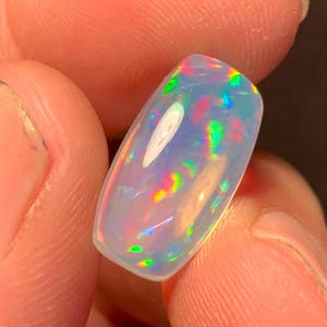 May include: A polished, oval-shaped opal gemstone. The opal displays a milky white base color with iridescent flashes of green, red, and blue. The stone is held between fingers, showcasing its vibrant play-of-color.